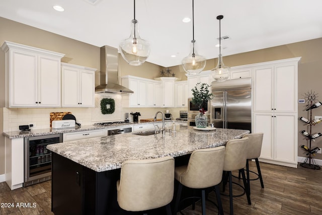 kitchen with an island with sink, appliances with stainless steel finishes, wine cooler, pendant lighting, and wall chimney exhaust hood