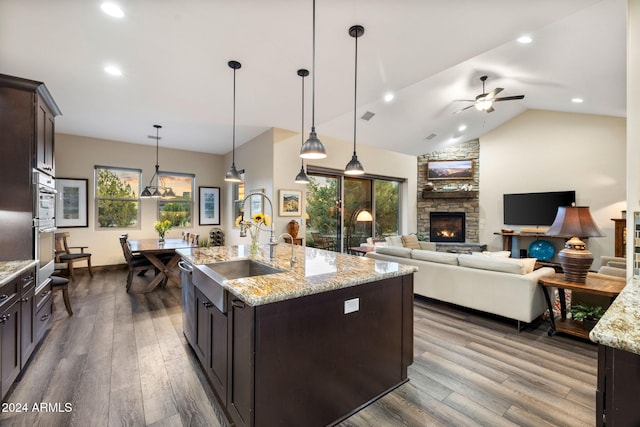 kitchen featuring wood-type flooring, hanging light fixtures, dark brown cabinetry, light stone counters, and a center island with sink