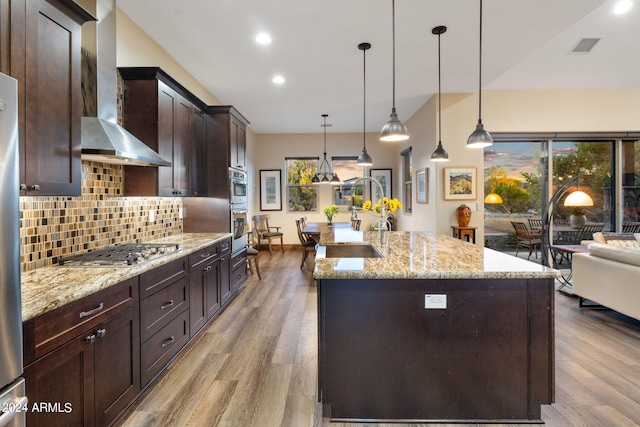 kitchen featuring wall chimney range hood, a center island with sink, appliances with stainless steel finishes, light hardwood / wood-style floors, and decorative light fixtures