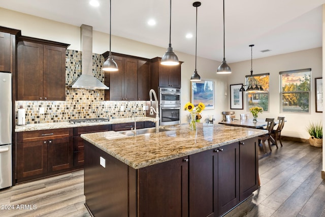 kitchen with wall chimney range hood, an island with sink, wood-type flooring, decorative light fixtures, and appliances with stainless steel finishes