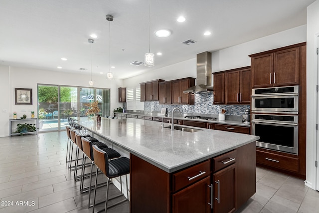 kitchen with decorative light fixtures, light stone countertops, stainless steel appliances, wall chimney exhaust hood, and an island with sink