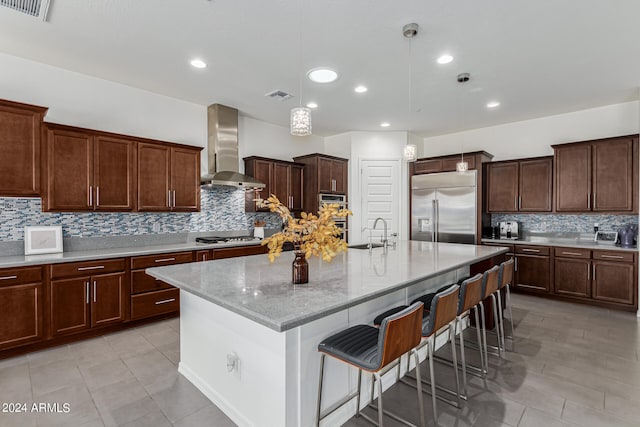 kitchen with stainless steel appliances, light stone counters, an island with sink, hanging light fixtures, and wall chimney exhaust hood