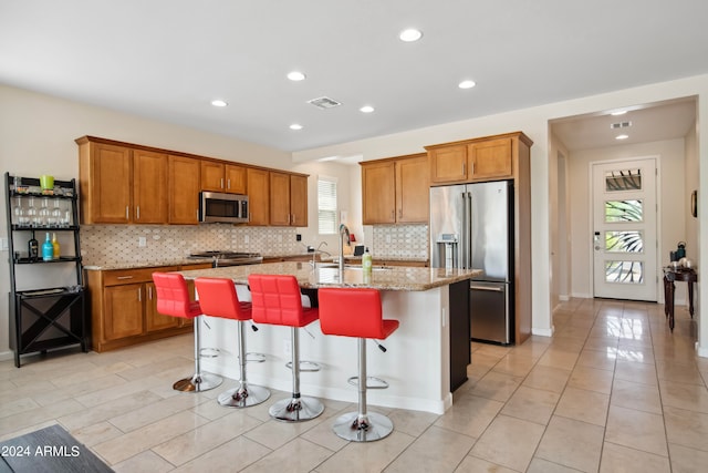 kitchen featuring tasteful backsplash, light tile patterned floors, light stone counters, a kitchen island with sink, and appliances with stainless steel finishes