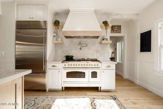 kitchen featuring custom exhaust hood, premium appliances, tasteful backsplash, light wood-type flooring, and white cabinets