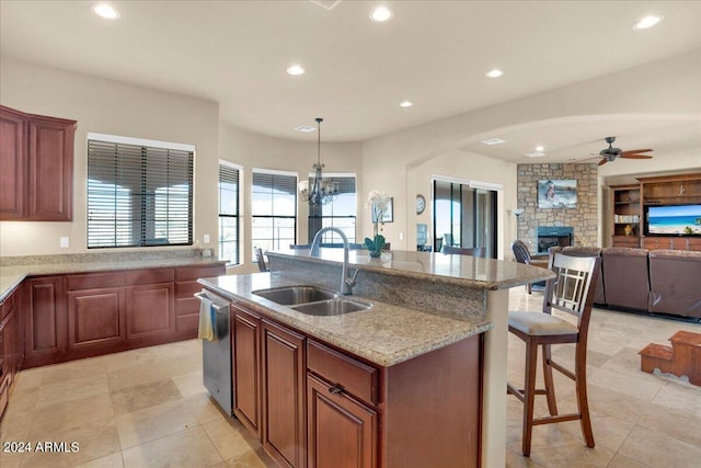 kitchen featuring light stone counters, a kitchen island with sink, a fireplace, ceiling fan with notable chandelier, and sink