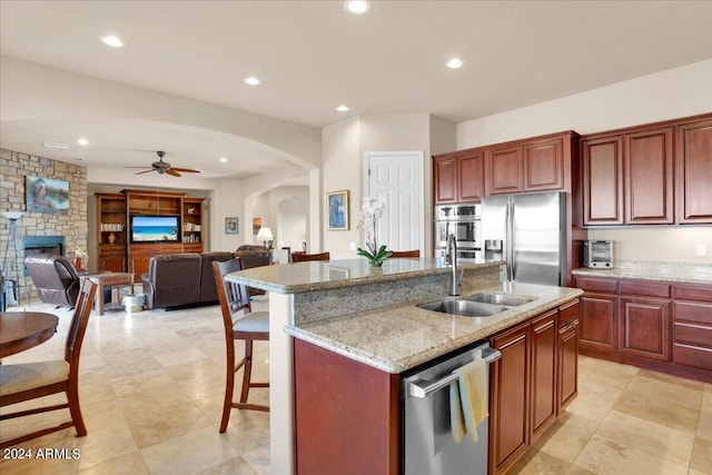 kitchen with ceiling fan, a kitchen island with sink, sink, stainless steel appliances, and a stone fireplace