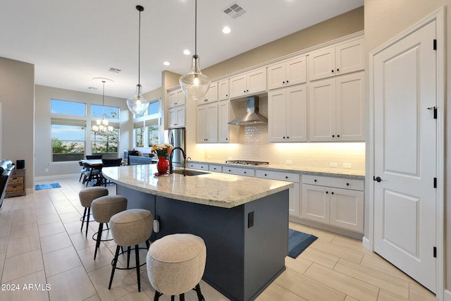 kitchen featuring light stone counters, an island with sink, tasteful backsplash, wall chimney range hood, and white cabinets