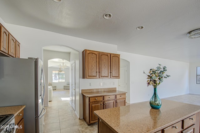 kitchen with arched walkways, light tile patterned floors, a textured ceiling, freestanding refrigerator, and brown cabinets