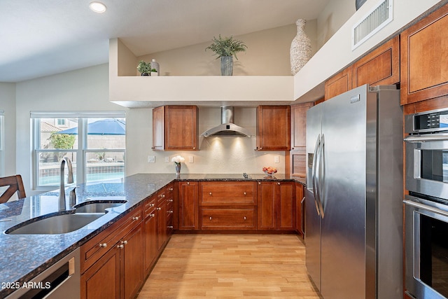 kitchen featuring stainless steel appliances, a sink, visible vents, wall chimney range hood, and dark stone countertops