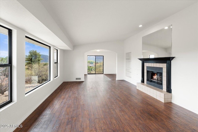 unfurnished living room with baseboards, built in features, arched walkways, a tiled fireplace, and wood finished floors