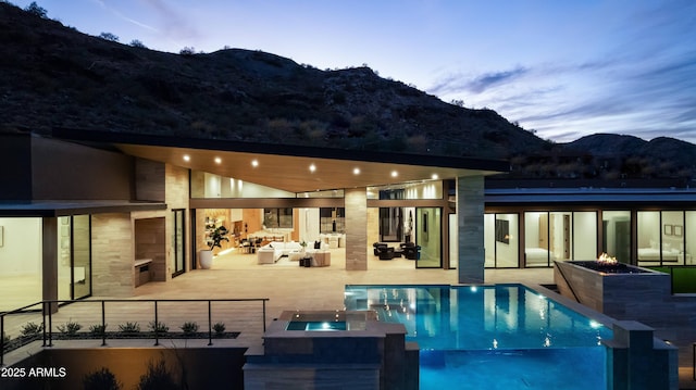 pool at dusk with a patio, a mountain view, an outdoor living space with a fireplace, and an in ground hot tub