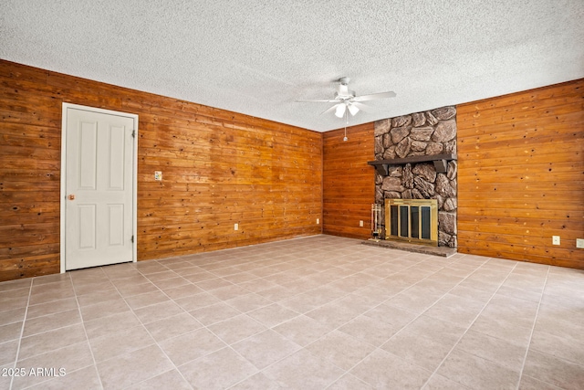 unfurnished living room with ceiling fan, a textured ceiling, a stone fireplace, wood walls, and light tile patterned floors