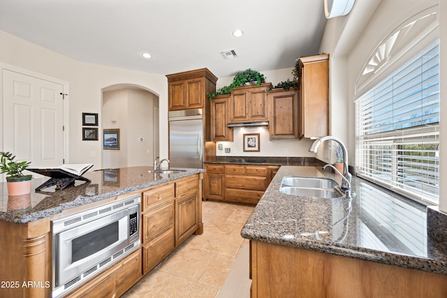 kitchen with built in appliances, sink, a kitchen island with sink, and dark stone counters