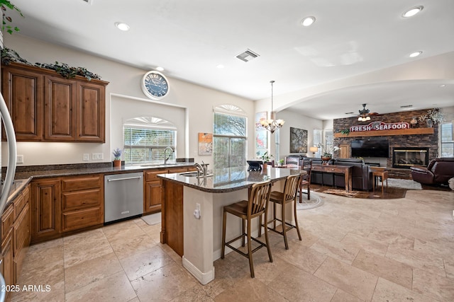 kitchen featuring a stone fireplace, ceiling fan with notable chandelier, sink, stainless steel dishwasher, and a center island with sink