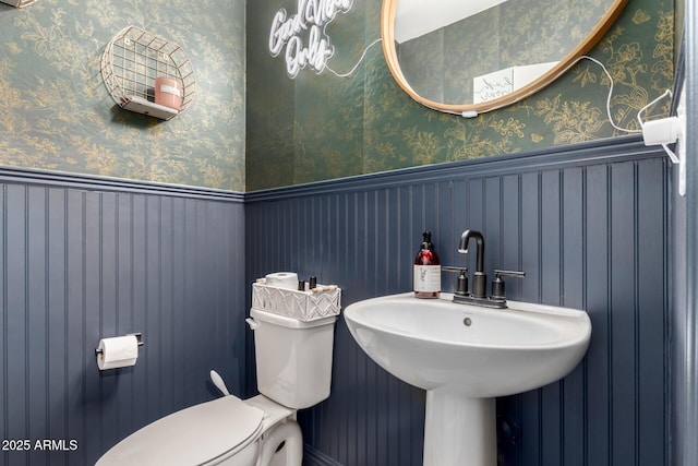 bathroom featuring a wainscoted wall, a sink, toilet, and wallpapered walls