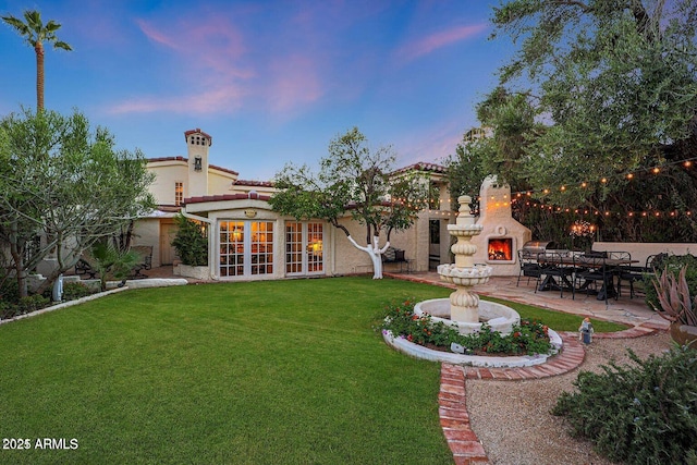 back house at dusk with french doors, exterior fireplace, a yard, and a patio