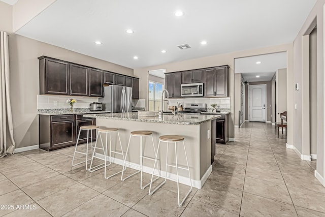 kitchen with dark brown cabinetry, a kitchen bar, visible vents, appliances with stainless steel finishes, and light stone counters