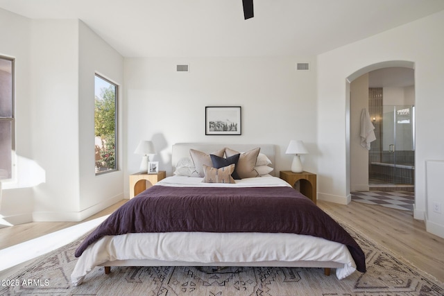 bedroom featuring wood finished floors, arched walkways, baseboards, ensuite bathroom, and a ceiling fan
