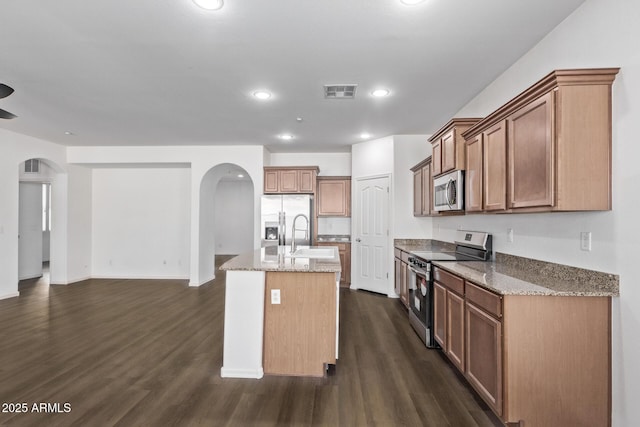 kitchen with sink, dark wood-type flooring, appliances with stainless steel finishes, light stone countertops, and an island with sink