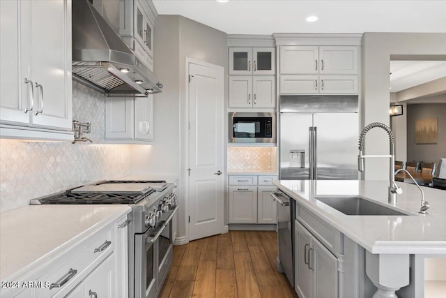 kitchen with tasteful backsplash, built in appliances, wood-type flooring, sink, and wall chimney range hood