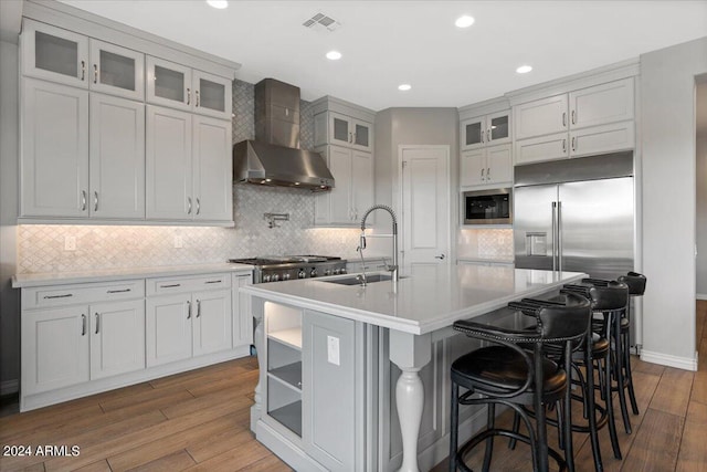 kitchen with an island with sink, sink, wall chimney range hood, hardwood / wood-style flooring, and a kitchen breakfast bar