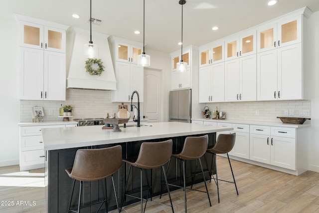 kitchen with white cabinetry, hanging light fixtures, a kitchen island with sink, stainless steel appliances, and custom range hood