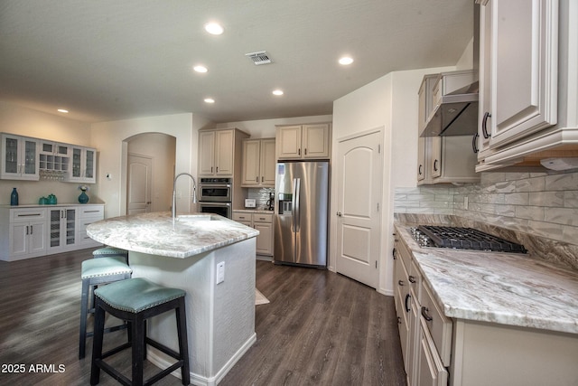 kitchen with stainless steel appliances, light stone countertops, a center island with sink, and backsplash