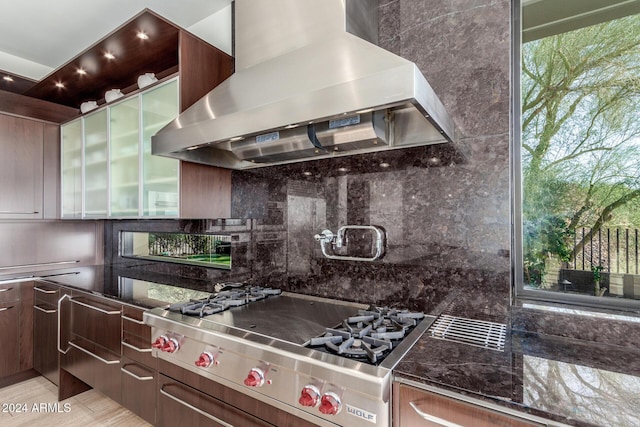 kitchen with dark stone counters, wall chimney range hood, decorative backsplash, dark brown cabinets, and stainless steel gas cooktop