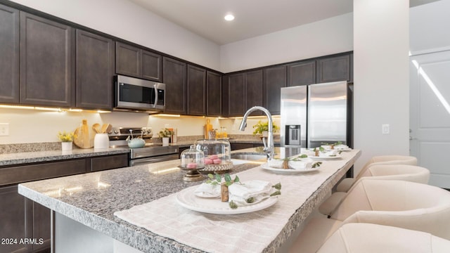 kitchen with a kitchen bar, an island with sink, a sink, appliances with stainless steel finishes, and dark brown cabinets