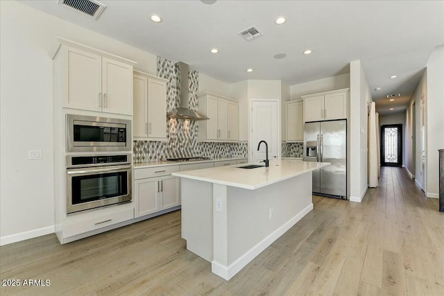 kitchen featuring wall chimney exhaust hood, sink, stainless steel appliances, a kitchen island with sink, and backsplash