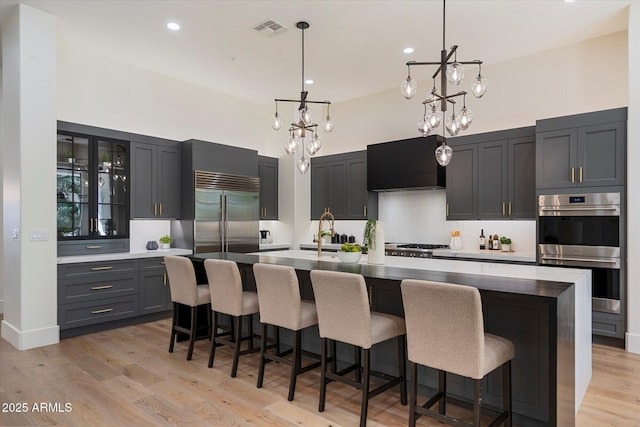 kitchen with a notable chandelier, appliances with stainless steel finishes, light wood finished floors, extractor fan, and glass insert cabinets