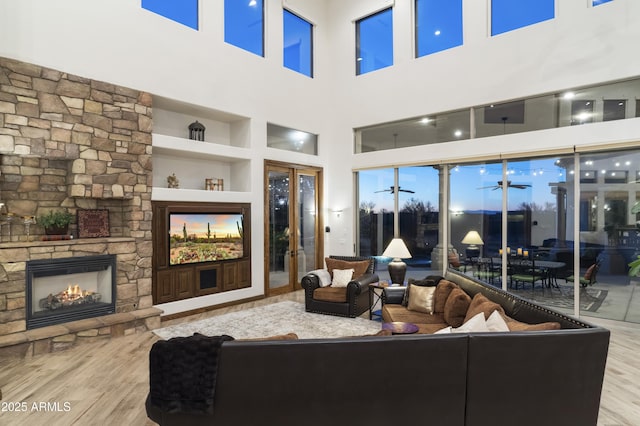 living room featuring a stone fireplace, light hardwood / wood-style flooring, built in features, ceiling fan, and a high ceiling