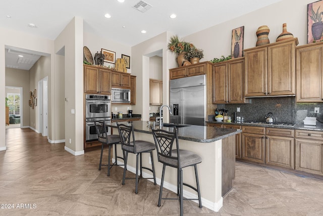 kitchen featuring built in appliances, a center island with sink, a breakfast bar, dark stone counters, and backsplash