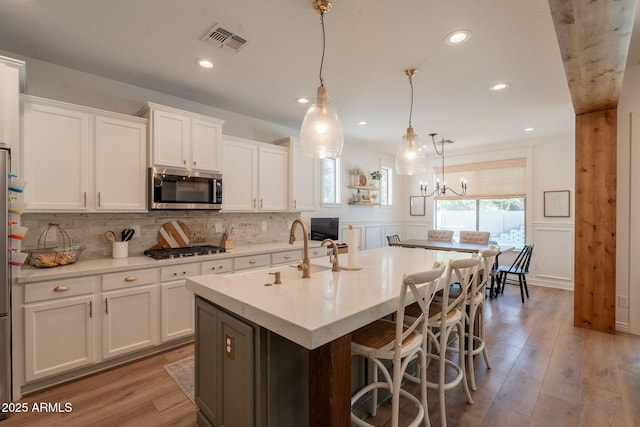 kitchen featuring stainless steel microwave, white cabinets, a kitchen island with sink, a decorative wall, and wood finished floors