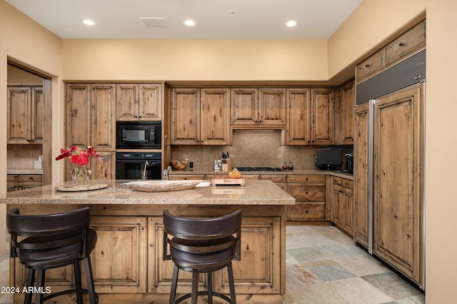 kitchen with black appliances, a kitchen breakfast bar, an island with sink, tasteful backsplash, and light stone counters