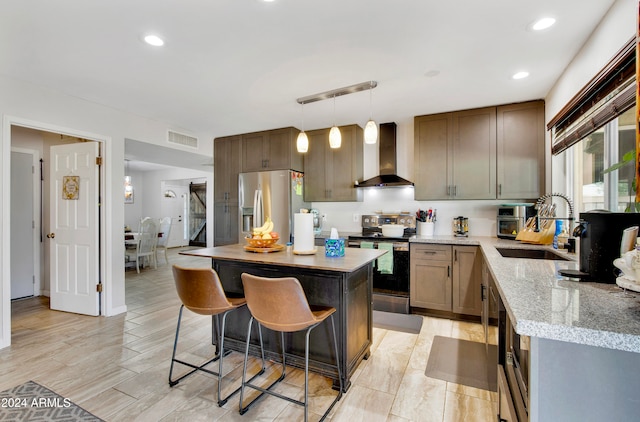 kitchen with wall chimney range hood, hanging light fixtures, appliances with stainless steel finishes, sink, and a center island