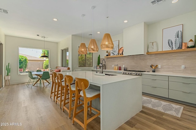 kitchen with sink, backsplash, an island with sink, decorative light fixtures, and light wood-type flooring