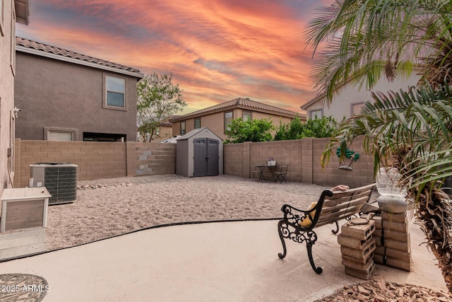 patio terrace at dusk featuring cooling unit, a storage unit, a fenced backyard, and an outdoor structure