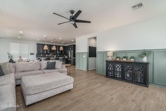 living room with a wainscoted wall, light wood-style flooring, visible vents, and ceiling fan