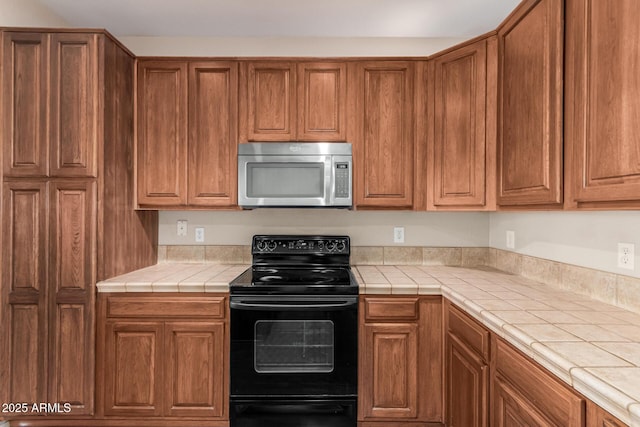 kitchen featuring tile counters, brown cabinetry, stainless steel microwave, and black electric range oven