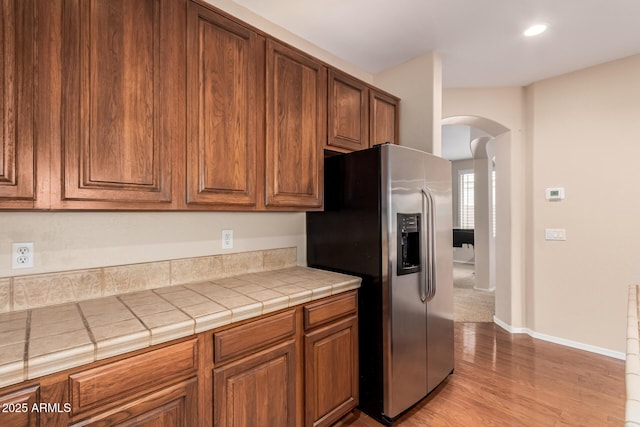 kitchen featuring arched walkways, light wood-style flooring, tile counters, brown cabinets, and stainless steel fridge