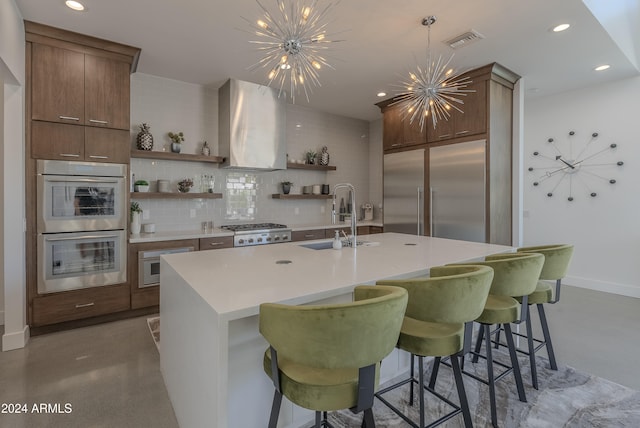 kitchen featuring wall chimney range hood, appliances with stainless steel finishes, a notable chandelier, and a kitchen island with sink