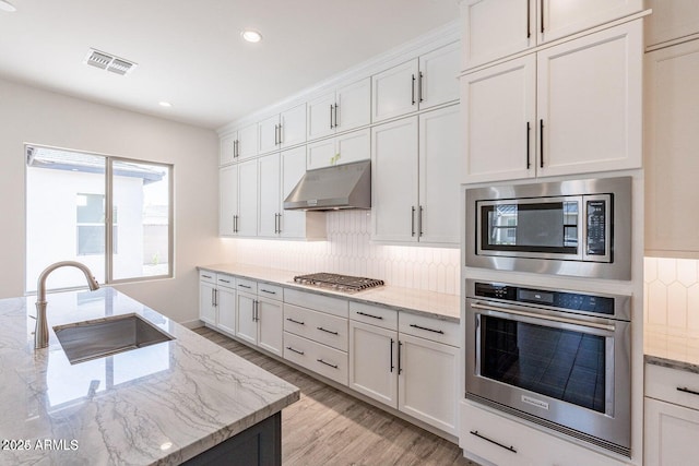 kitchen with backsplash, under cabinet range hood, appliances with stainless steel finishes, light wood-style floors, and a sink