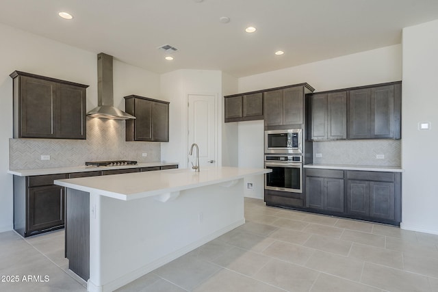 kitchen with dark brown cabinetry, sink, appliances with stainless steel finishes, a kitchen island with sink, and wall chimney range hood