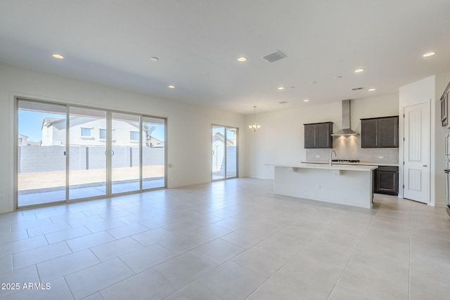 kitchen featuring light tile patterned flooring, an inviting chandelier, a center island with sink, wall chimney range hood, and backsplash