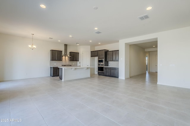 kitchen featuring appliances with stainless steel finishes, backsplash, a notable chandelier, an island with sink, and wall chimney exhaust hood