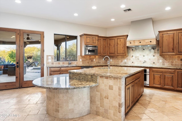 kitchen featuring sink, light stone counters, custom range hood, ceiling fan, and a kitchen island with sink