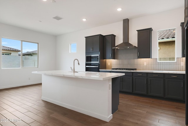 kitchen with stainless steel appliances, wall chimney exhaust hood, dark hardwood / wood-style flooring, a kitchen island with sink, and tasteful backsplash