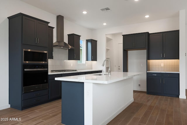 kitchen with a center island with sink, sink, stainless steel appliances, wall chimney exhaust hood, and tasteful backsplash