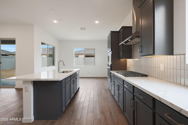 kitchen featuring a center island with sink, sink, wall chimney range hood, stainless steel appliances, and dark wood-type flooring